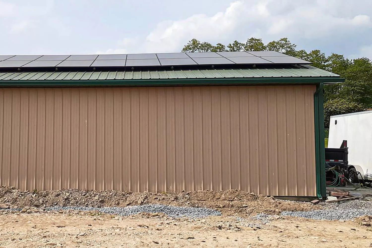 Tan metal shed with rooftop solar panels on a gravel lot, with trees in the background.
