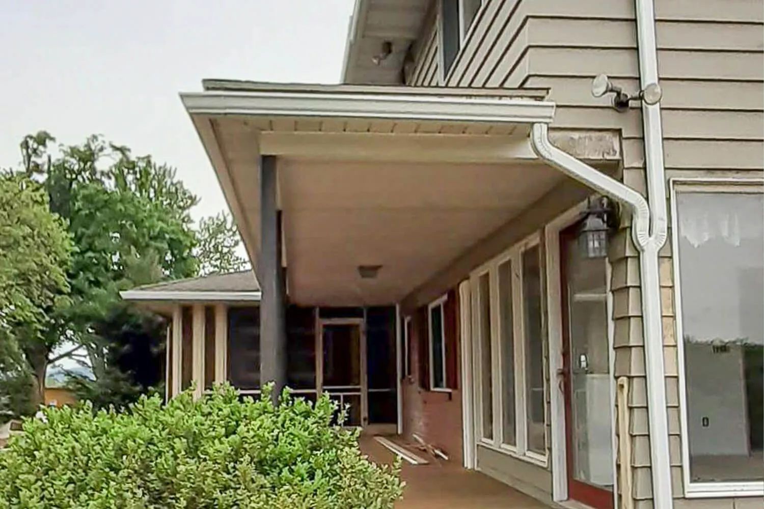 Covered apartment walkway with tan siding, white trim, and a porch lined by windows and bushes