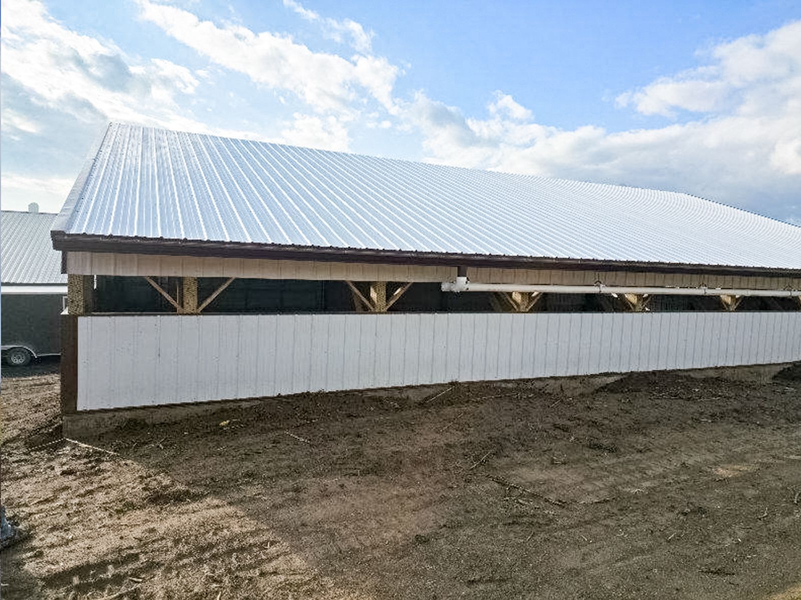 Long white metal-roofed barn behind a white fence on a muddy rural lot under a cloudy sky