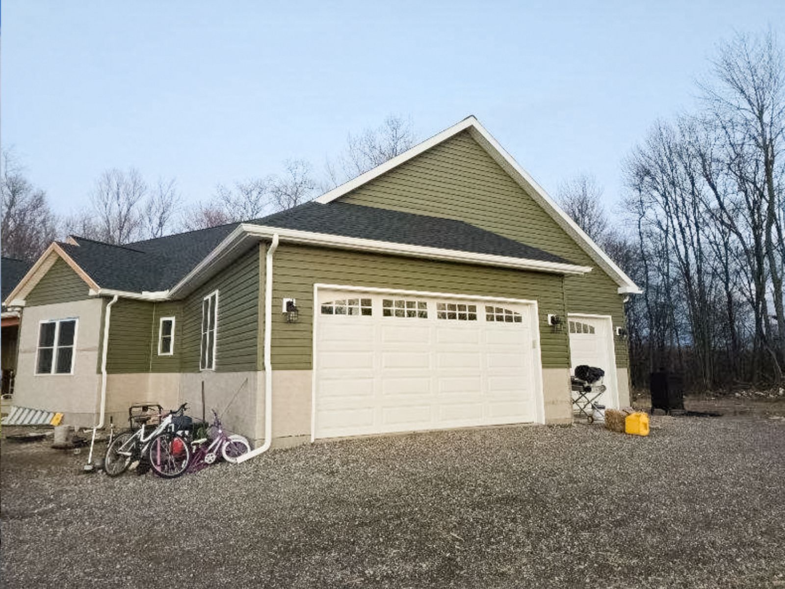 Green house with white garage door, bicycles by the driveway, and leafless trees in the background