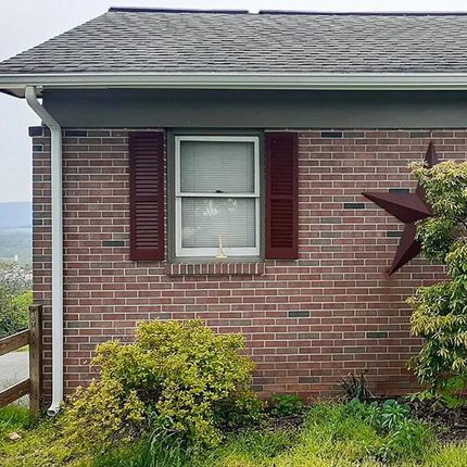 Brick house exterior with a white-framed window and dark shutters, plus bushes and a wooden railing.