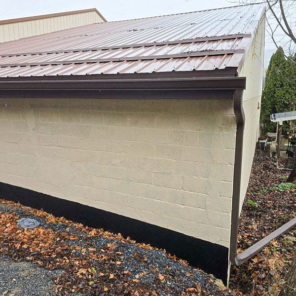Cream-colored building side with brown metal roof and black foundation trim beside a rocky path.