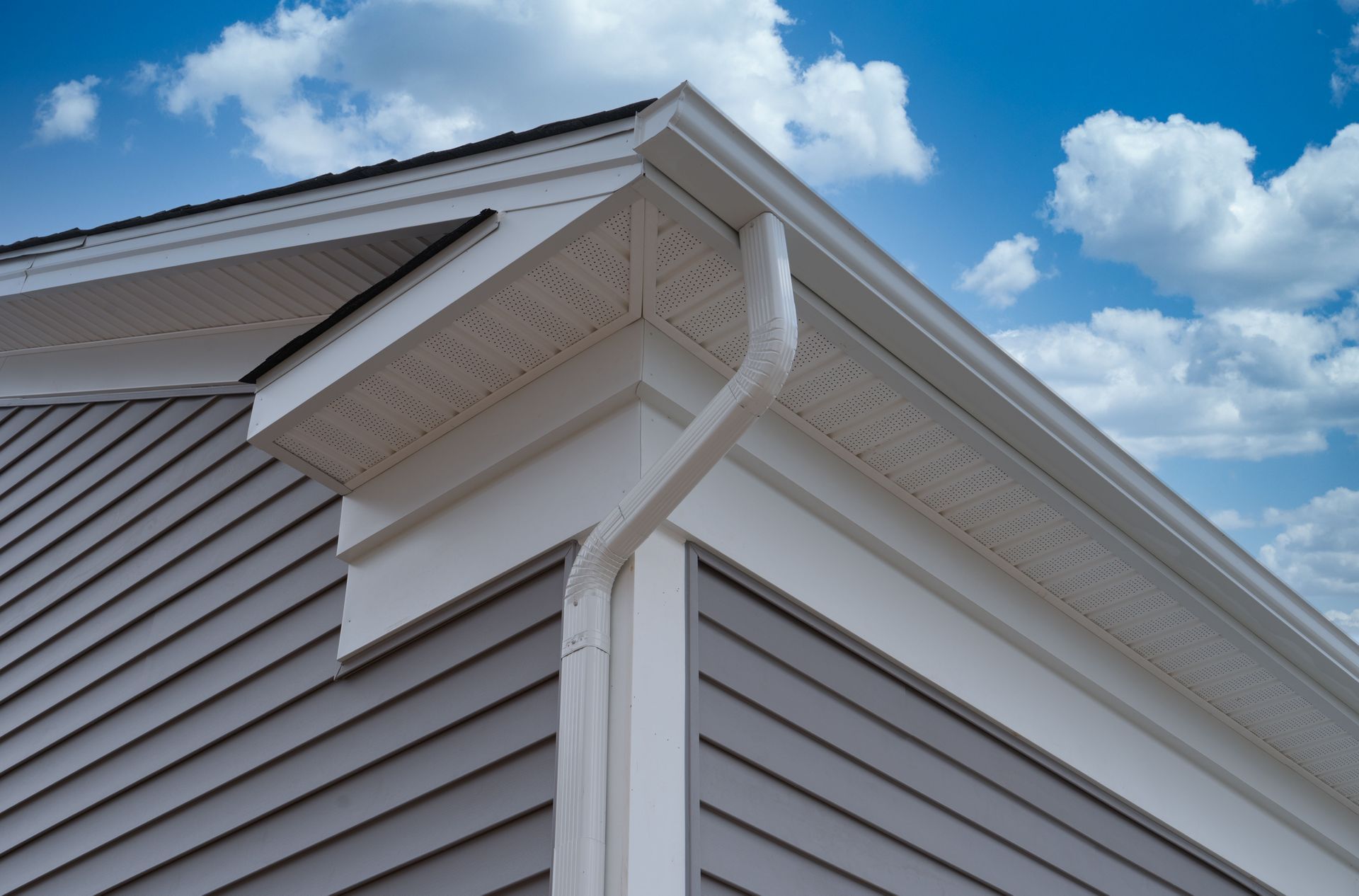 White house corner with vinyl siding, soffit vents, and a gutter against a blue sky with clouds