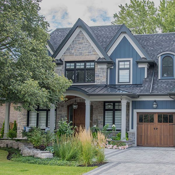 Modern two-story suburban house with dark blue siding, stone facade, and wood garage door, framed by trees and garden