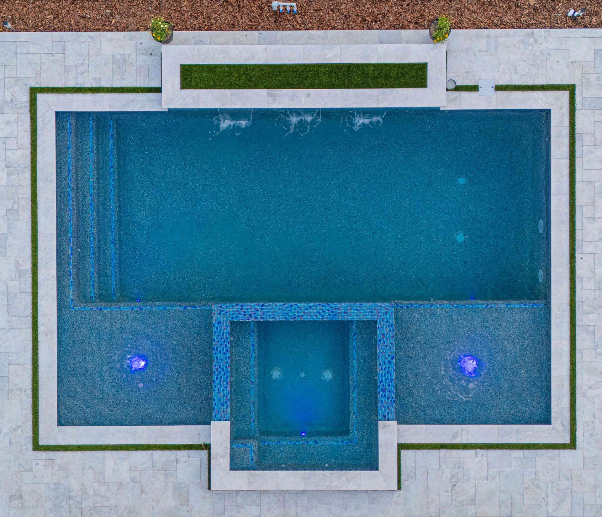 Aerial view of a rectangular backyard pool with a central spa and two glowing blue lights.