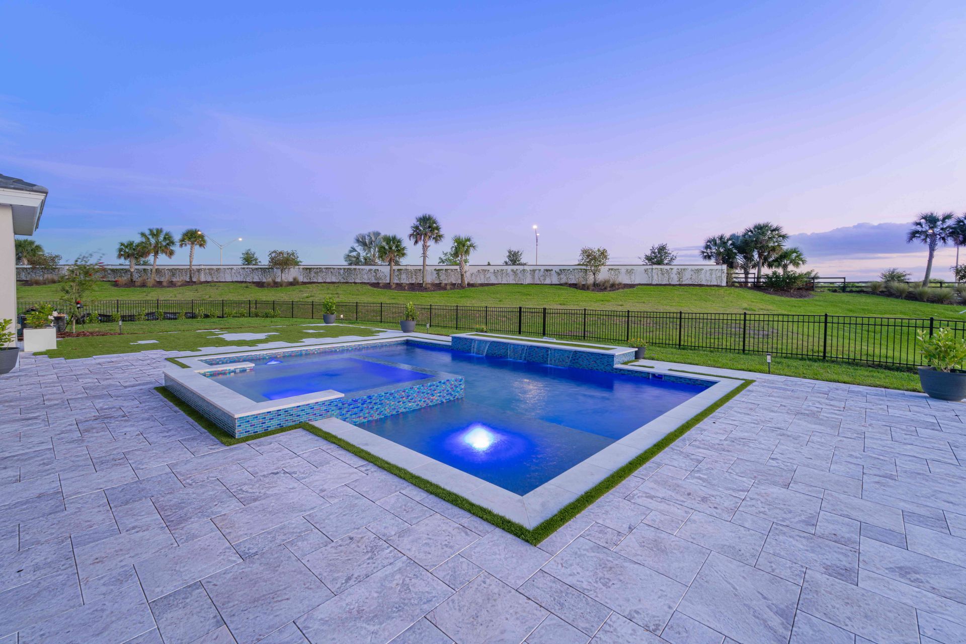 Modern patio with lit blue swimming pool, fenced lawn, and open sunset view over water