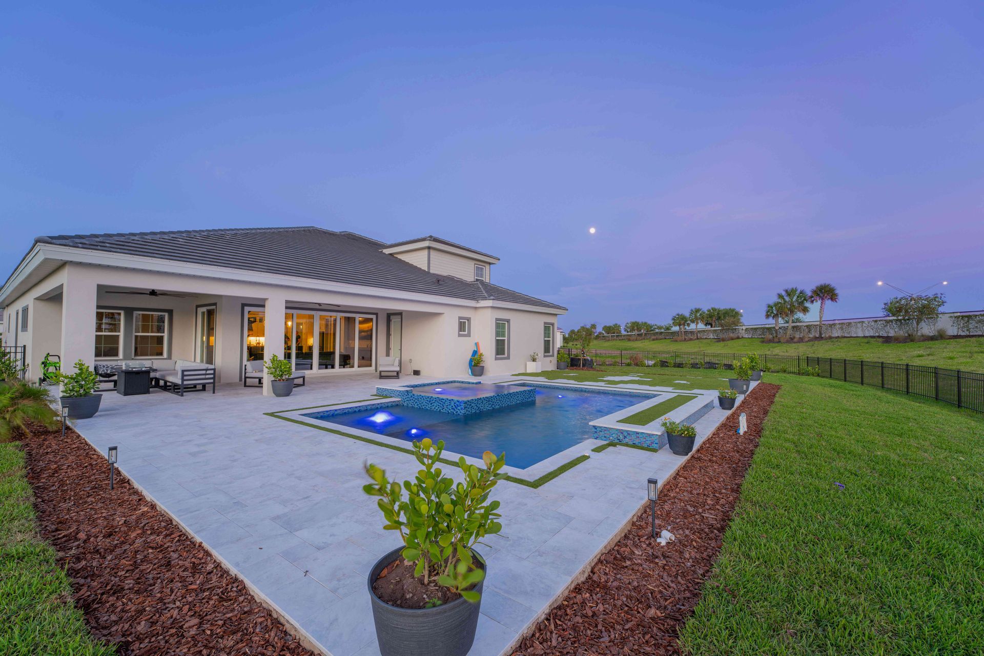 Modern house with lit patio and a blue swimming pool at dusk, bordered by a green lawn and landscaped yard