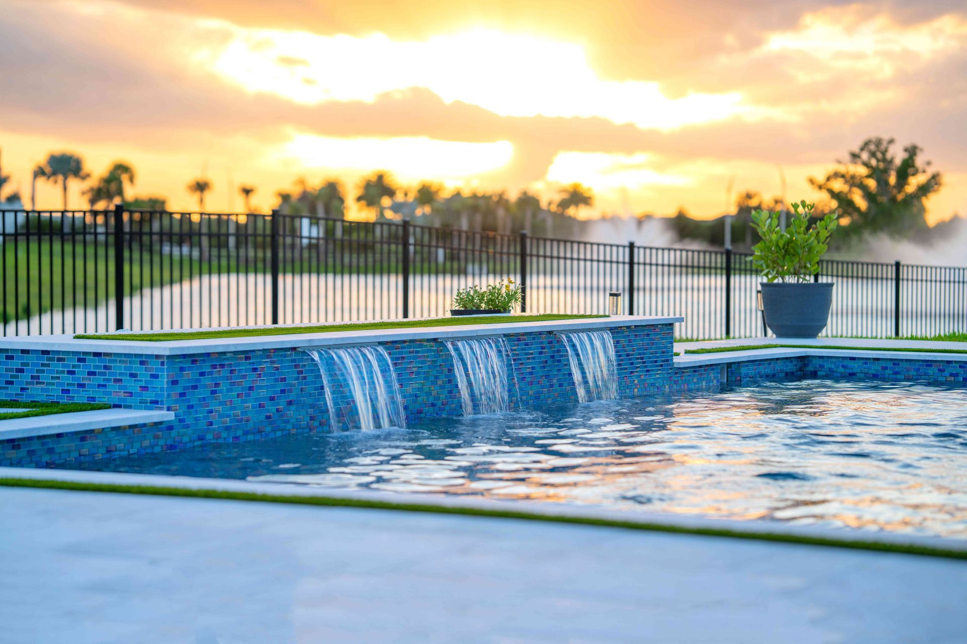 Backyard pool with a blue-tiled waterfall, reflecting sunset light behind a fence and potted plants