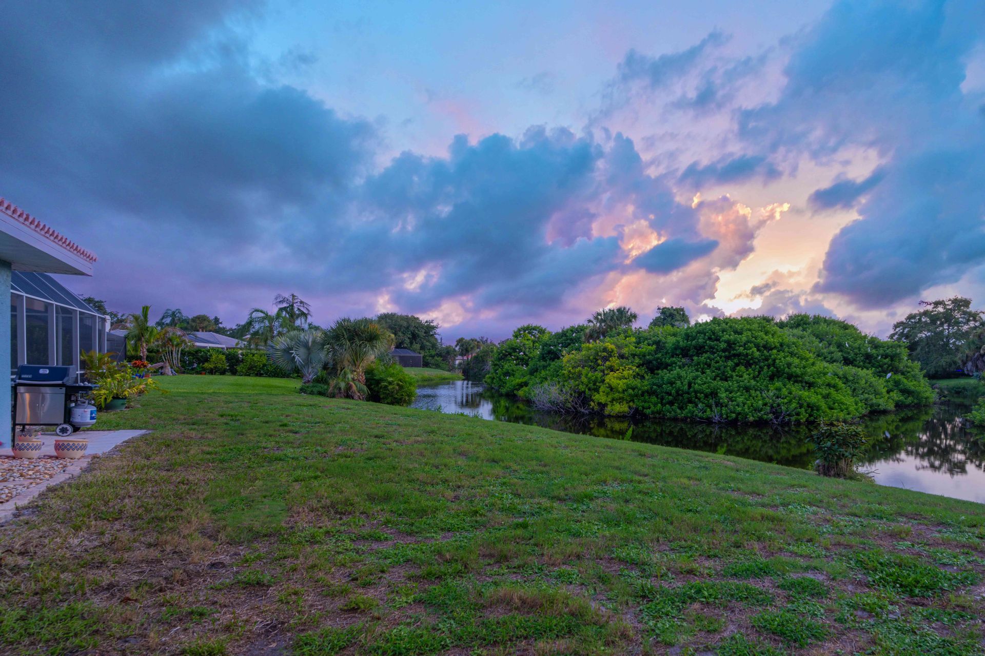 Grassy riverside lawn beside houses and trees under a colorful sunset sky