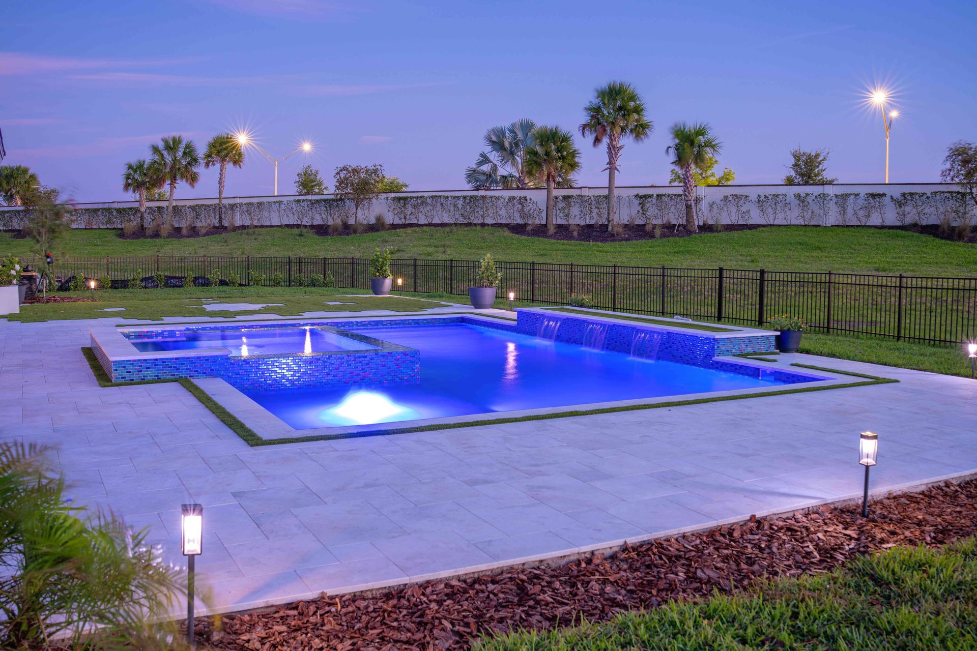 Illuminated outdoor swimming pool at dusk, bordered by hedges and palm trees with landscape lights.