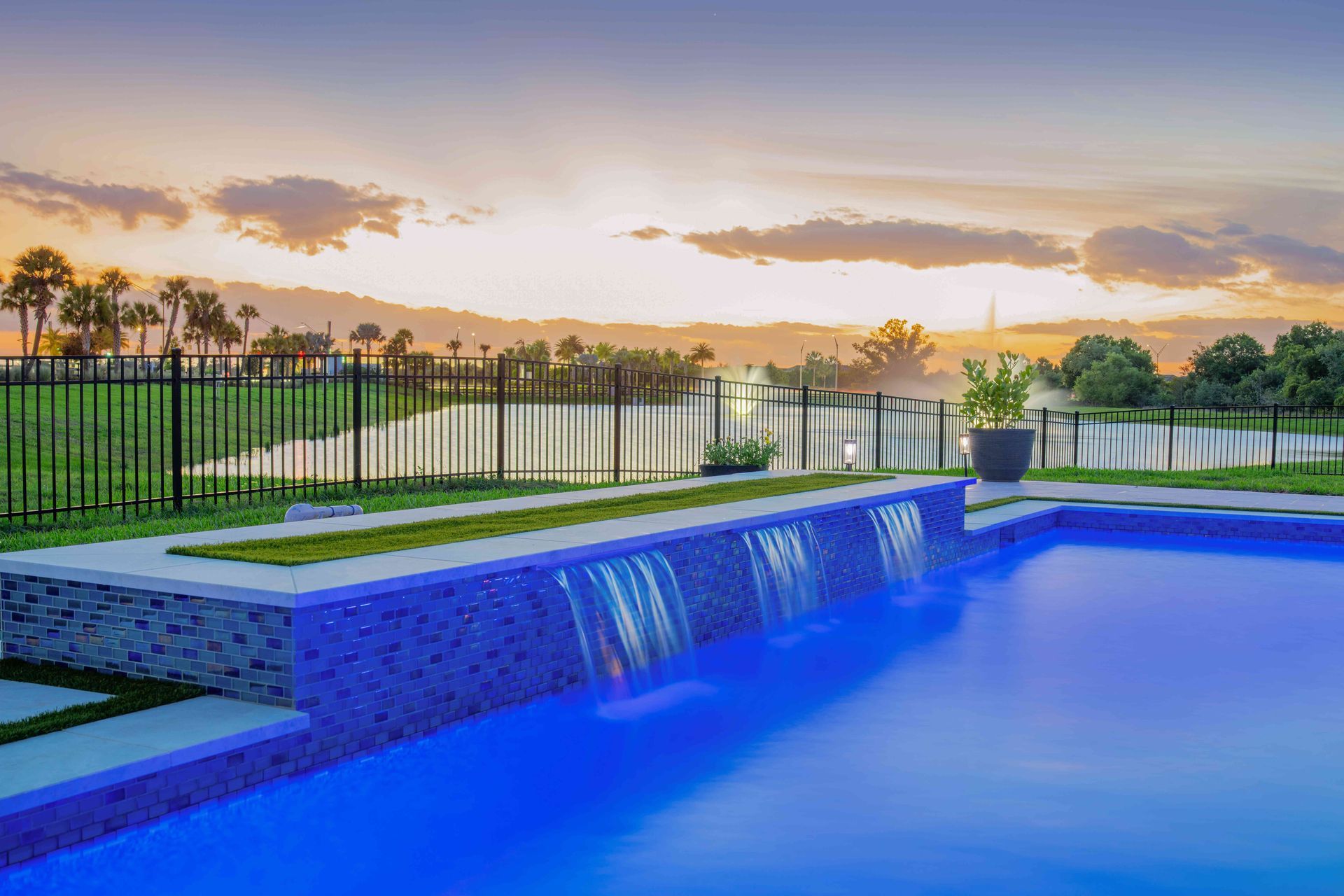 Modern blue pool with waterfall edge at sunset, bordered by a black fence and green lawn.