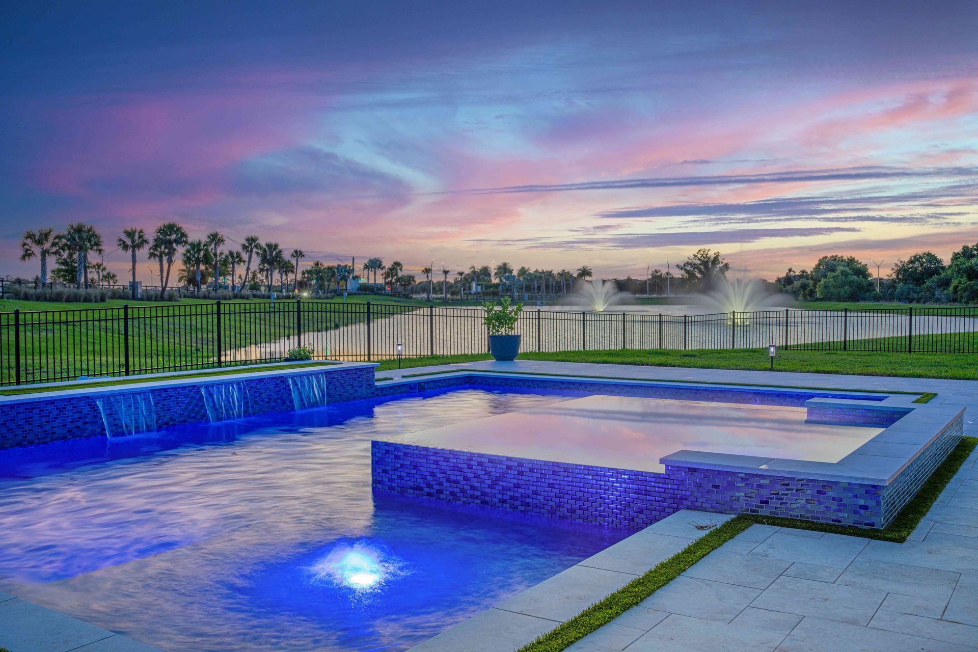 Modern pool with blue LED lighting and a sunset sky over a fenced patio.