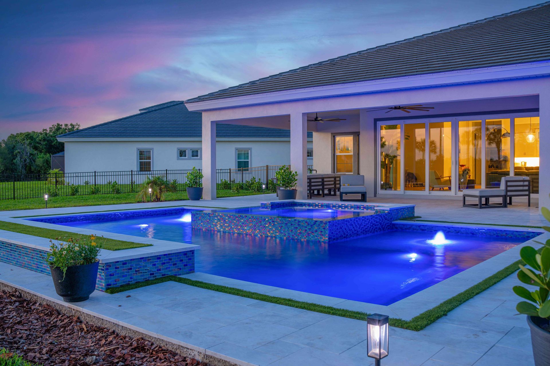 Modern house patio with glowing blue pool at dusk and warm lights through glass doors