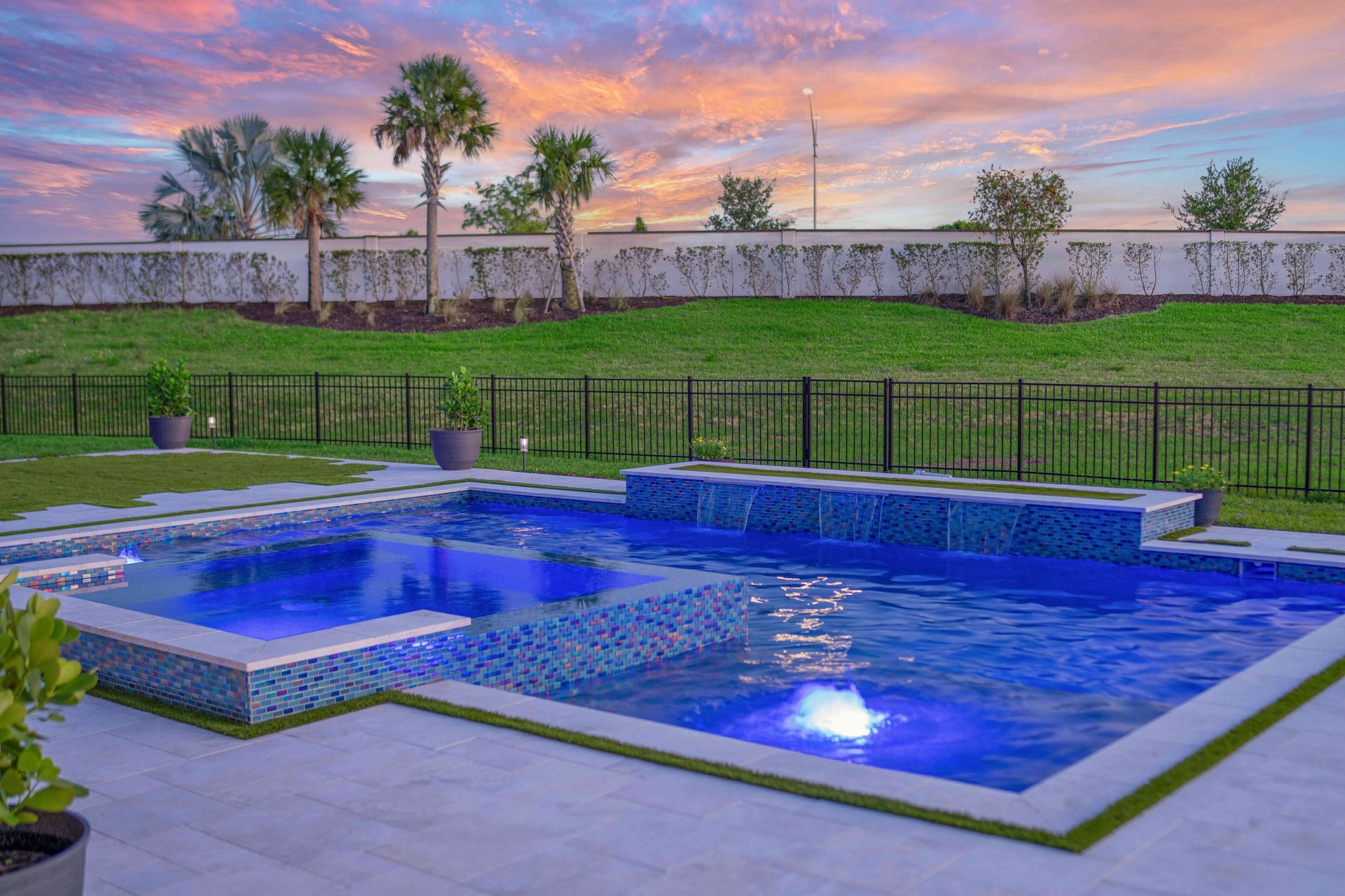 Blue-lit backyard pool at sunset with a landscaped lawn and distant fence.