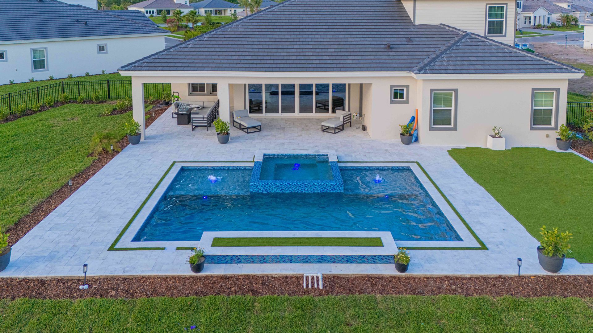 Modern house patio with a rectangular blue pool and hot tub, bordered by a light stone deck and green lawn.
