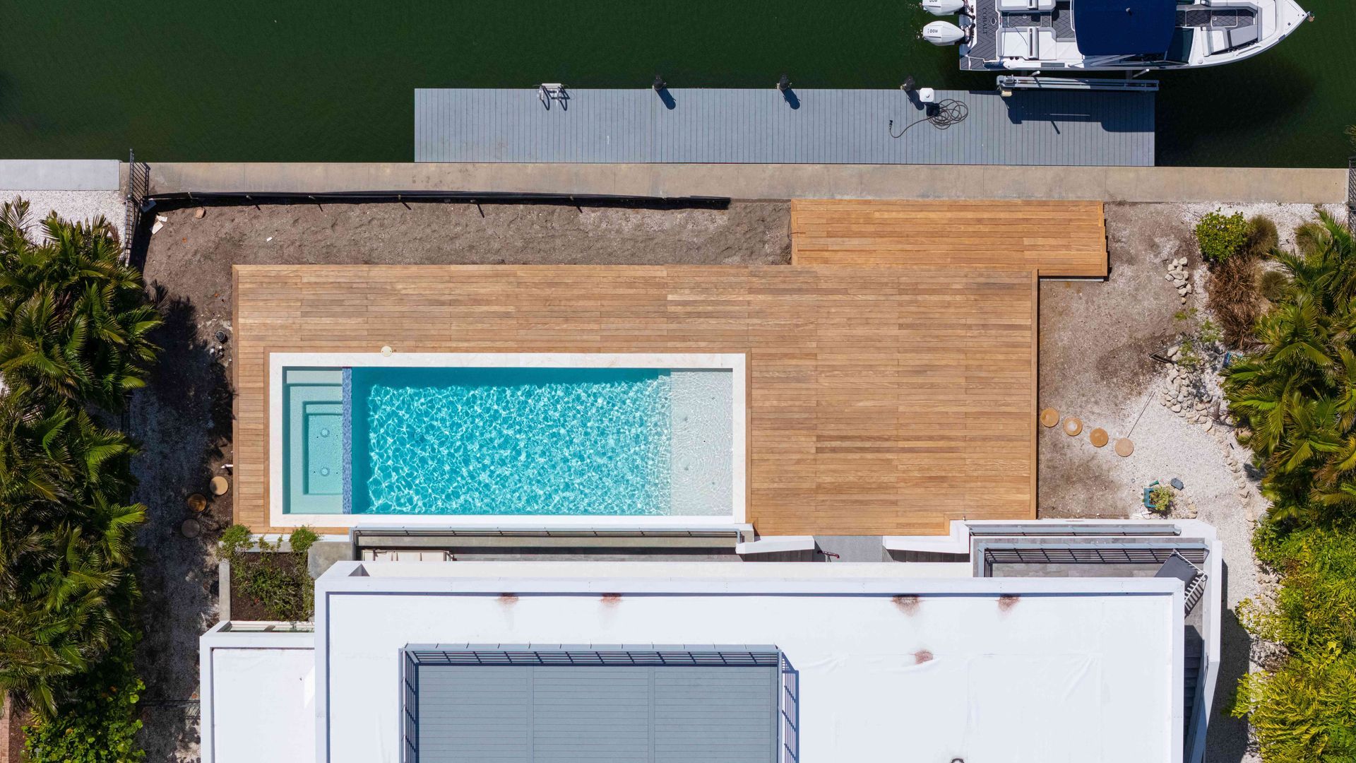 Aerial view of a rooftop deck with a turquoise pool beside a wooden patio and a boat dock nearby.
