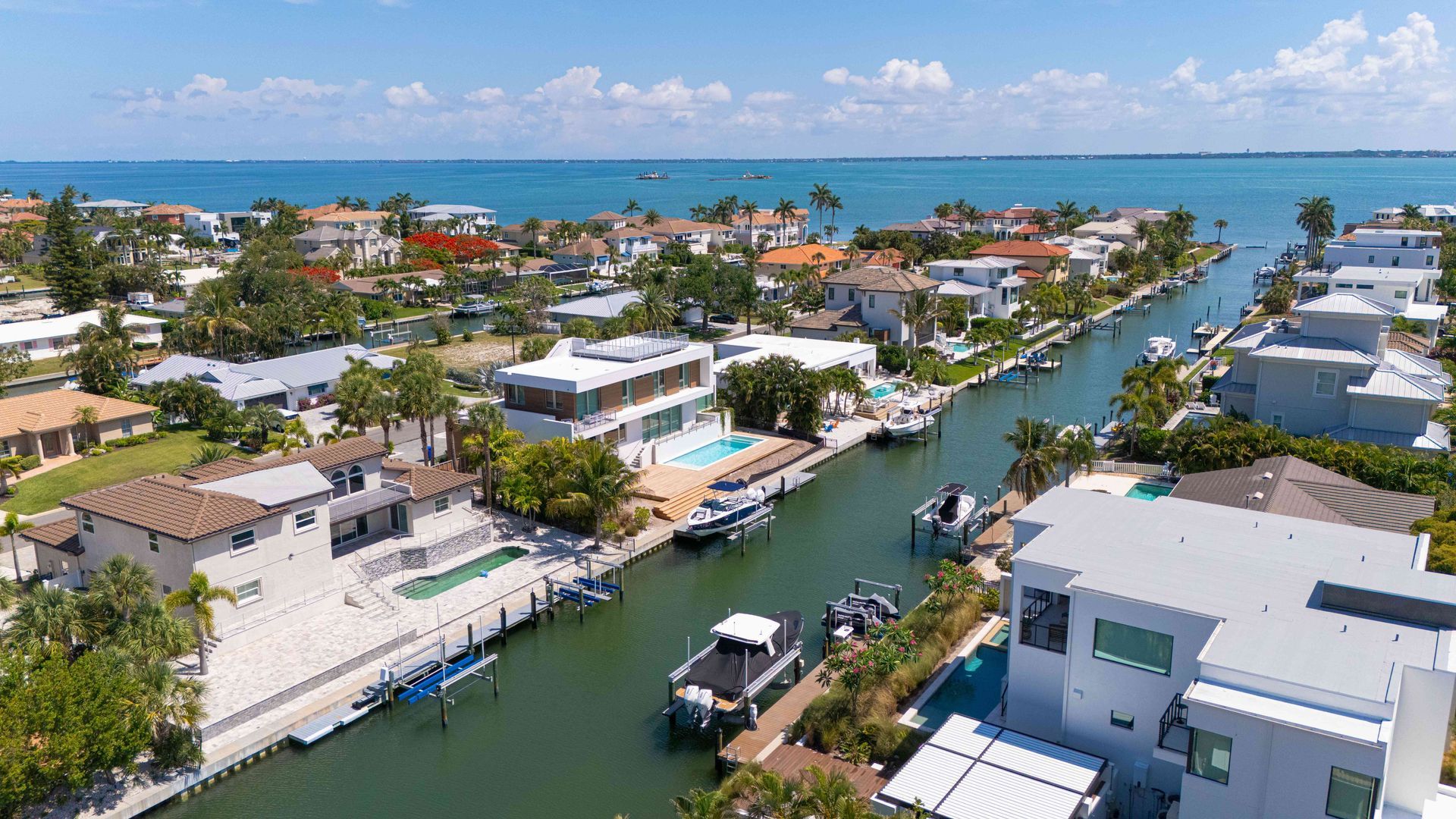 Aerial view of a sunny coastal canal lined with waterfront homes, boats, and palm trees.