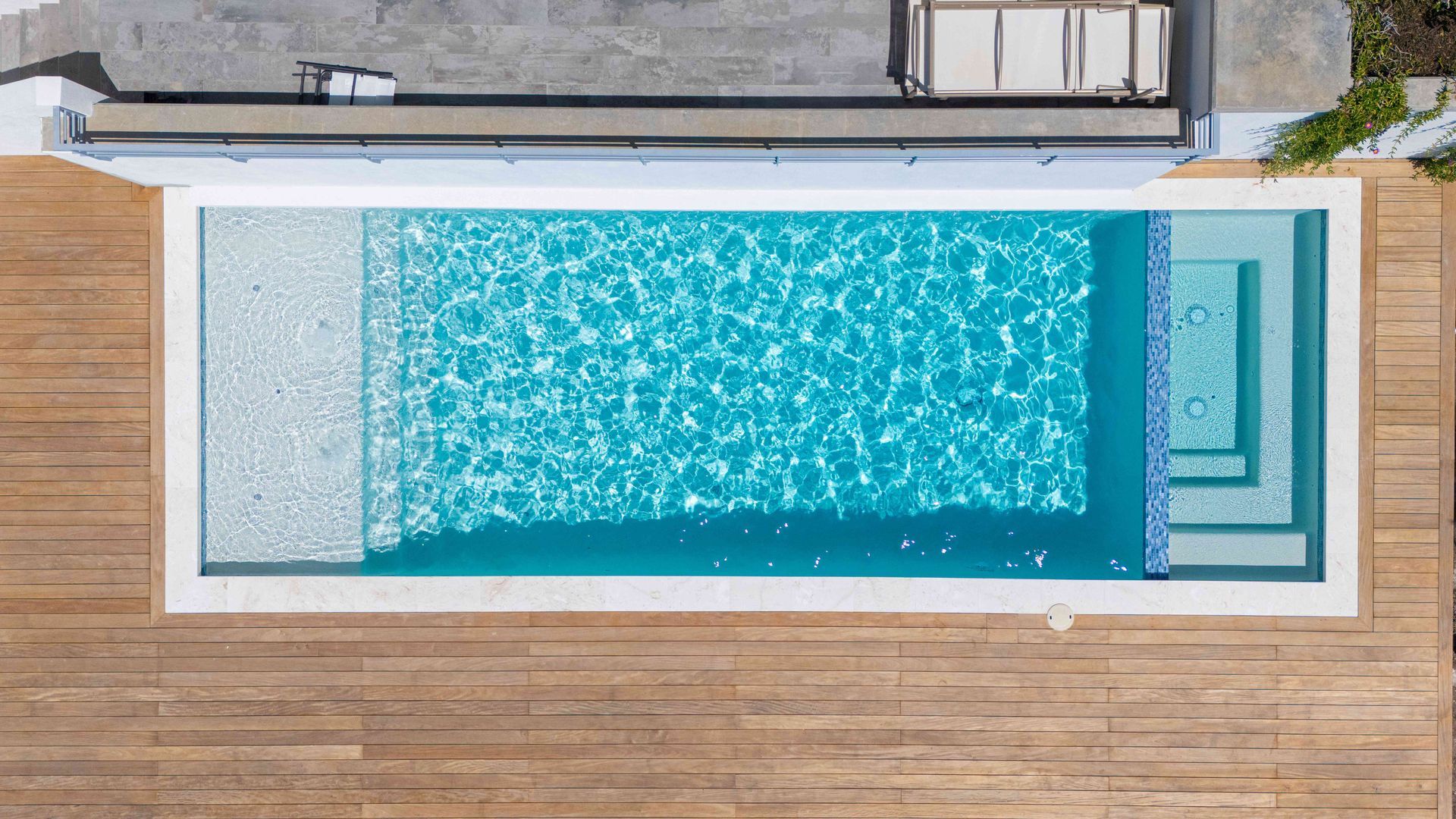 Overhead view of a rectangular turquoise swimming pool beside a wooden deck and concrete patio.