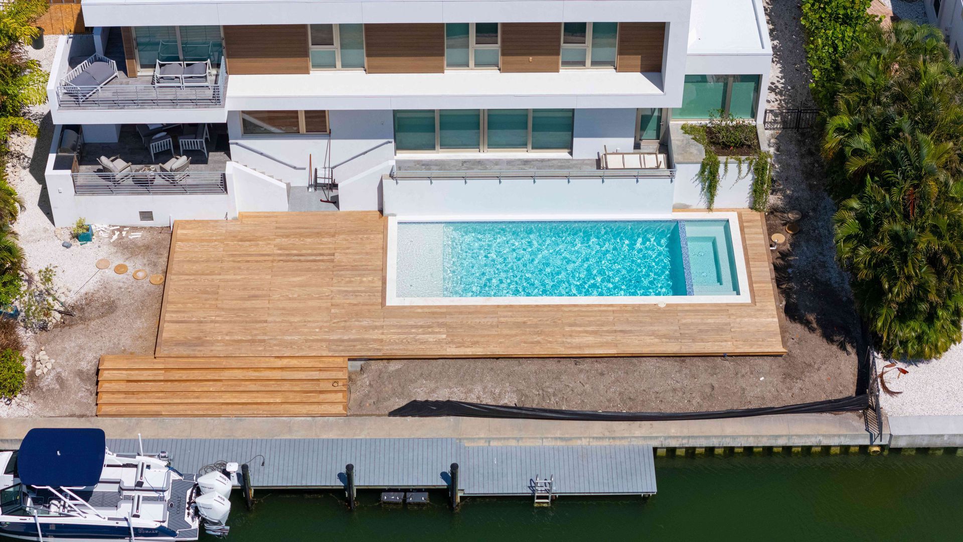 Aerial view of a modern waterfront home with a wooden deck, swimming pool, and docked boat.