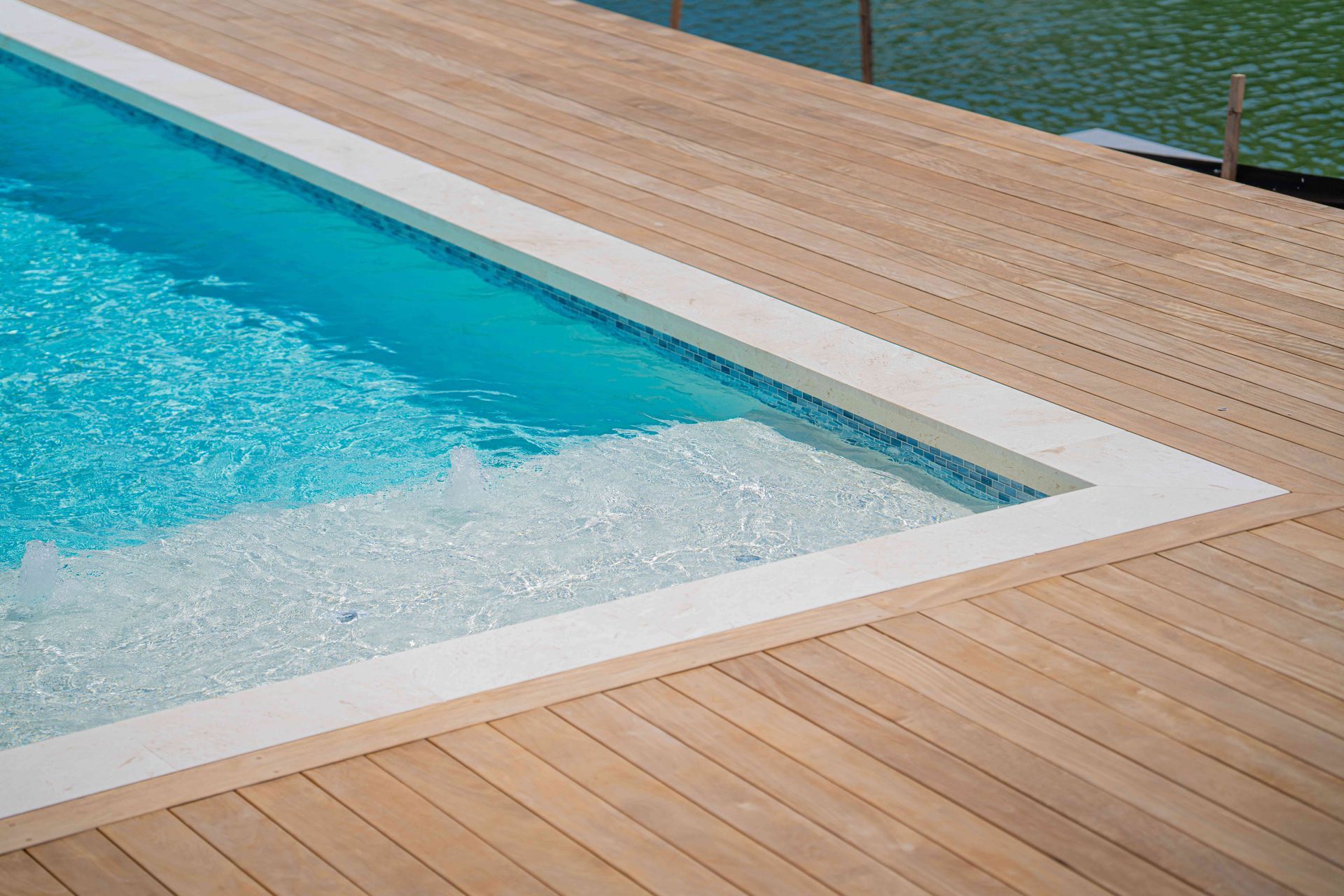 Corner of a turquoise swimming pool with white edging beside a light wood deck