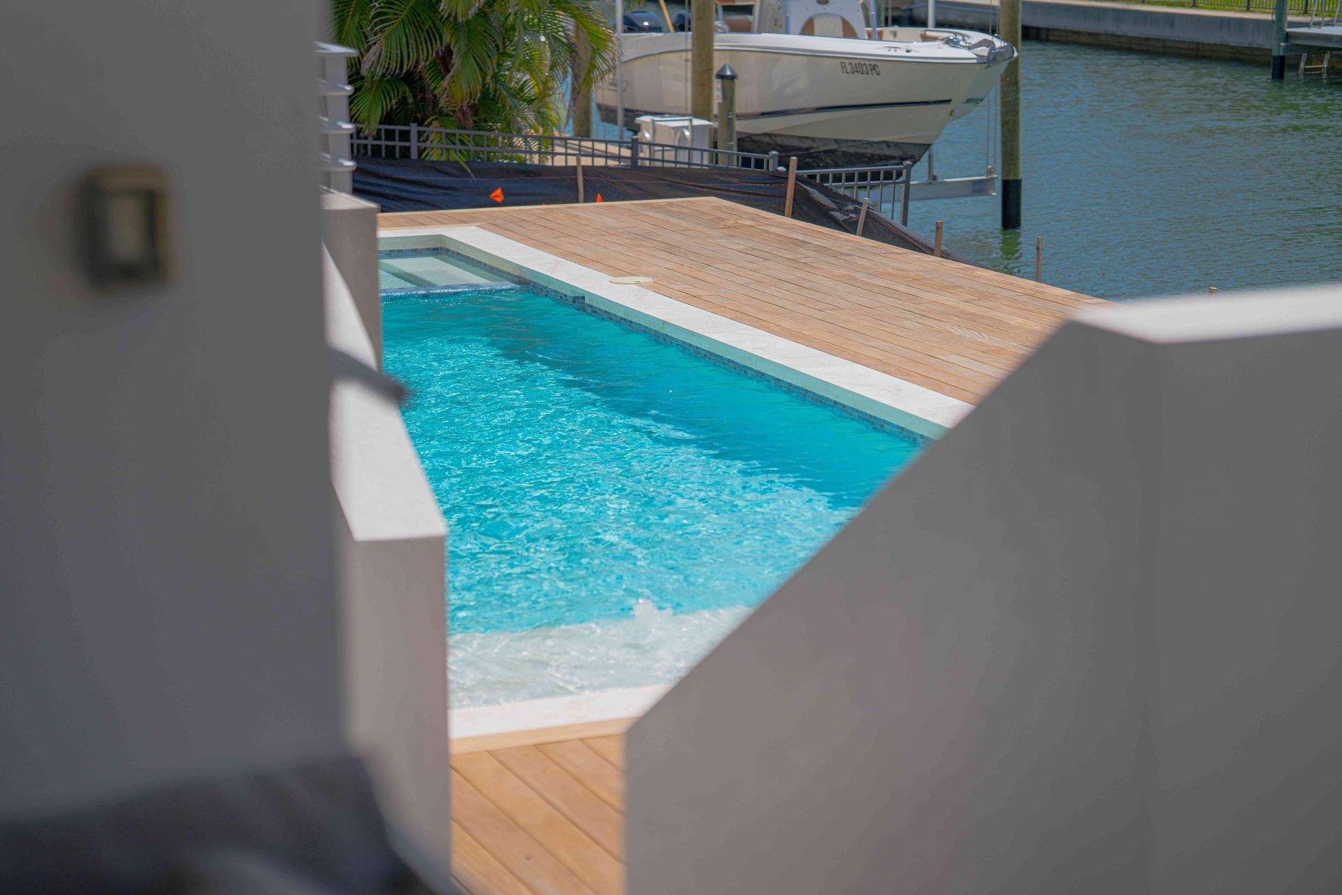 Bright turquoise pool beside a tan deck, viewed through white walls with water and boats in the background
