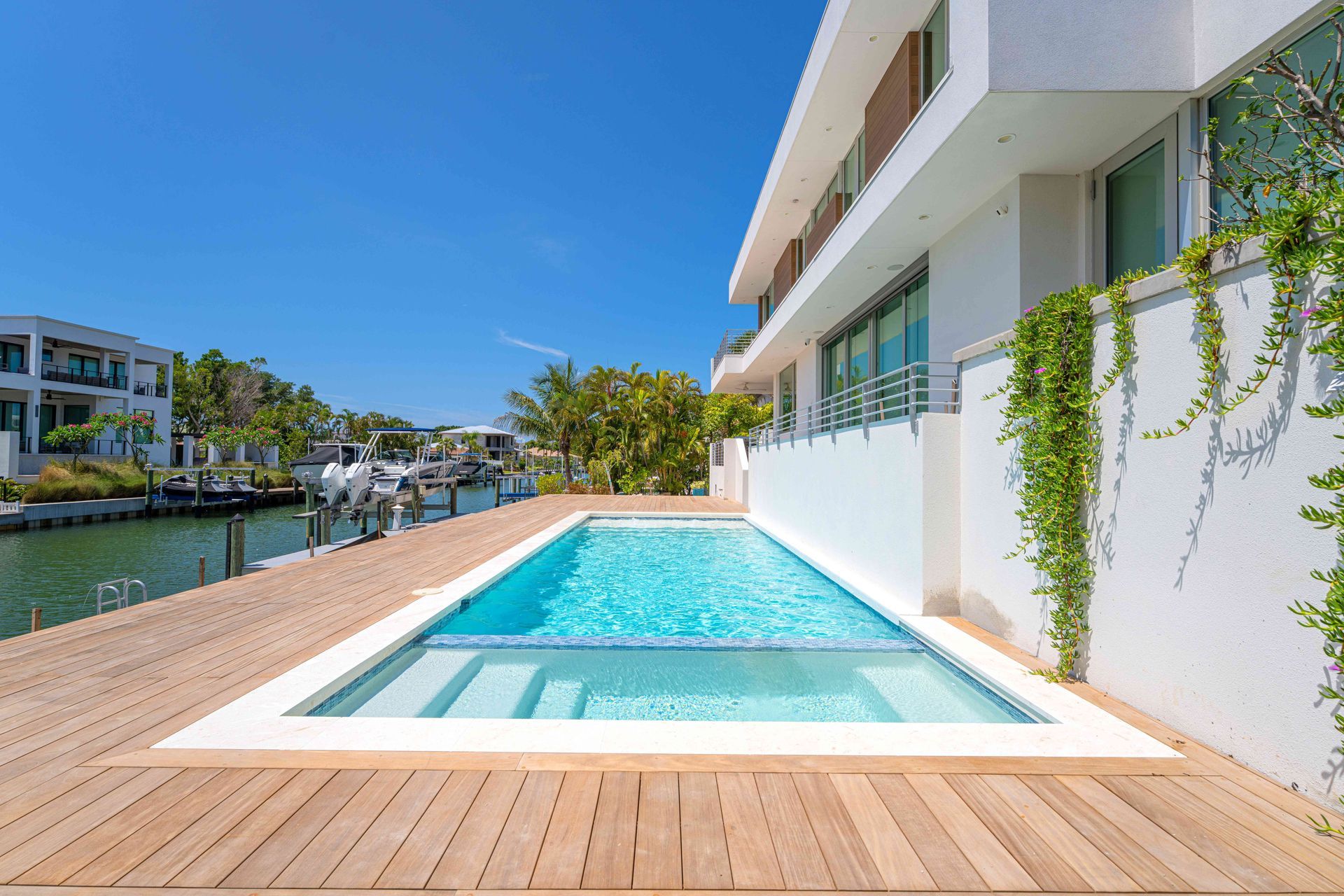 Modern waterfront patio with a small turquoise pool beside a white house and wooden deck