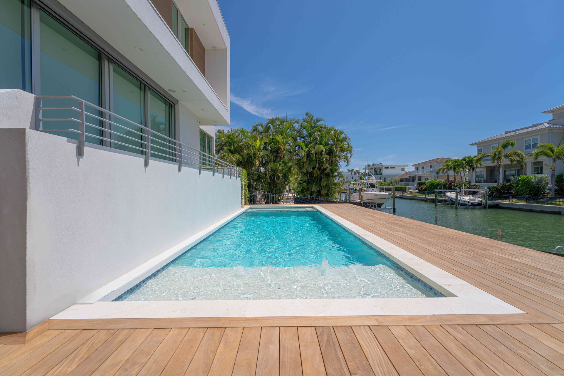 Modern backyard pool beside a white house, with a wooden deck and clear blue sky