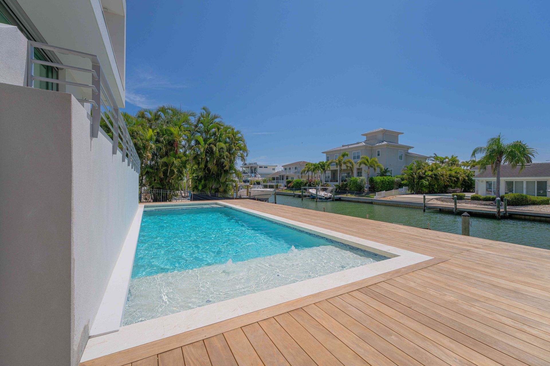 Modern backyard with a turquoise pool, wooden deck, and clear blue sky, bordered by white walls and greenery