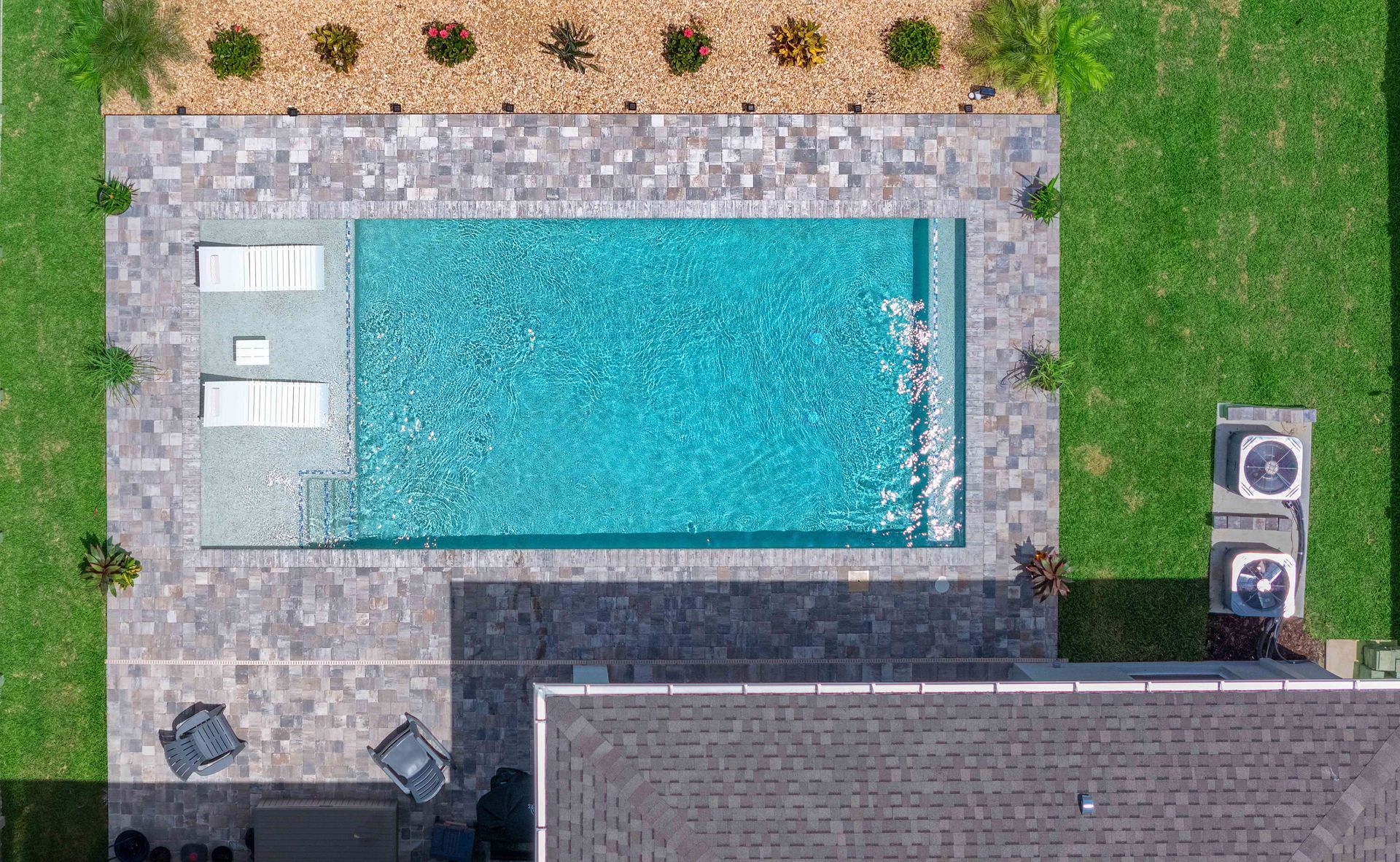 Aerial view of a backyard pool with lounge chairs, stone patio, grass, and a house roof edge