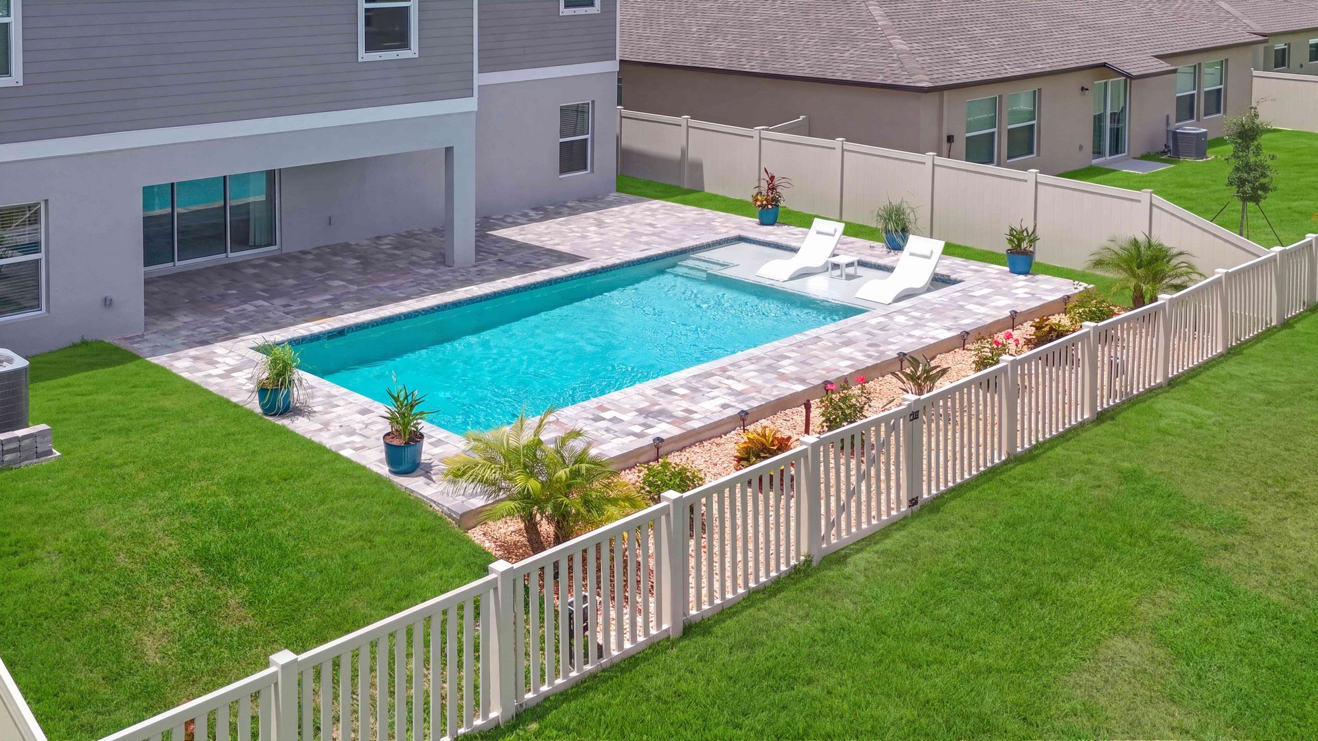 Backyard with a rectangular swimming pool, lounge chairs, and a white fence beside a house