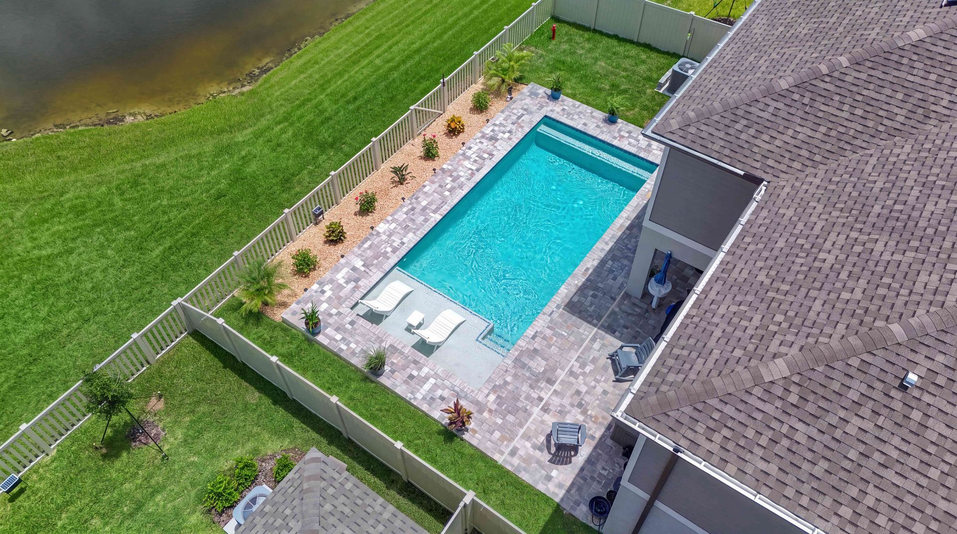 Aerial view of a backyard with a rectangular swimming pool, patio chairs, and a fenced lawn beside a house.
