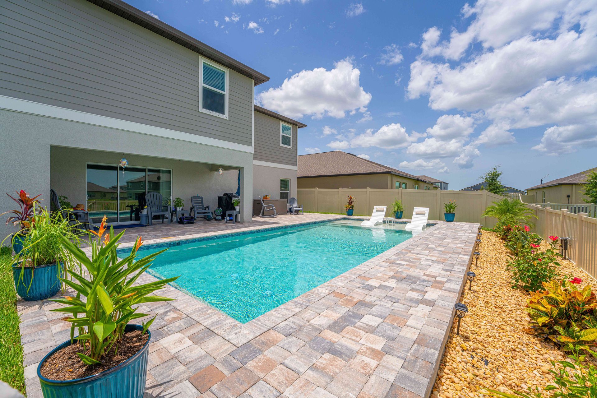 Backyard patio with a turquoise swimming pool beside a modern house and lounge chairs under a bright sky