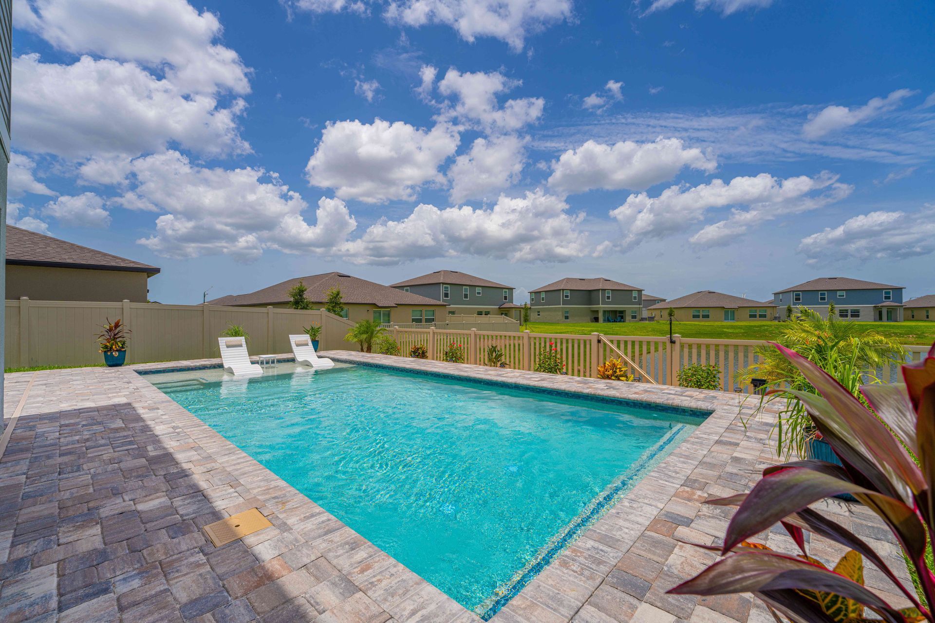 Backyard swimming pool with lounge chairs, stone patio, and cloudy blue sky over suburban homes