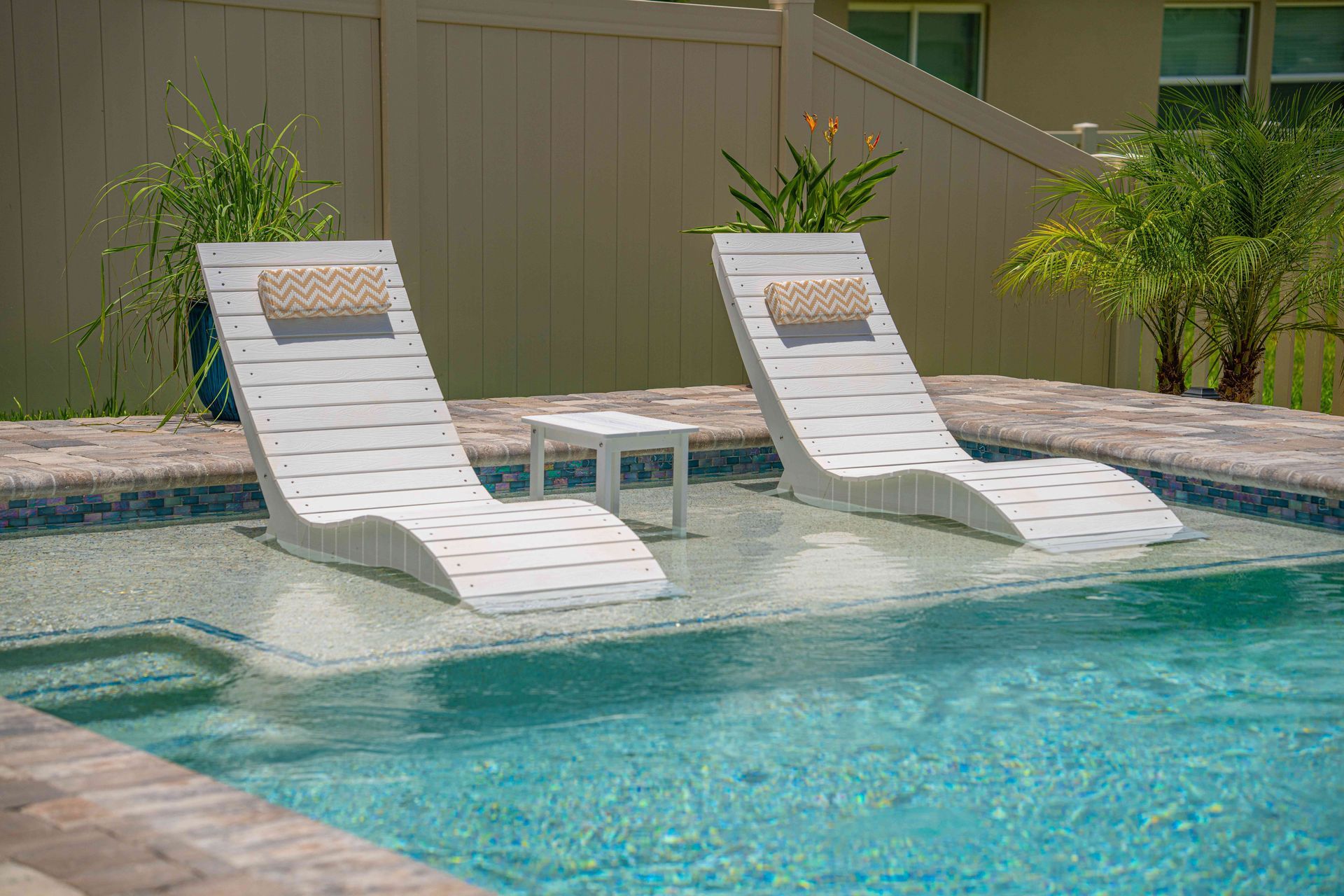 Two white lounge chairs beside a turquoise pool with a small table and tropical plants.