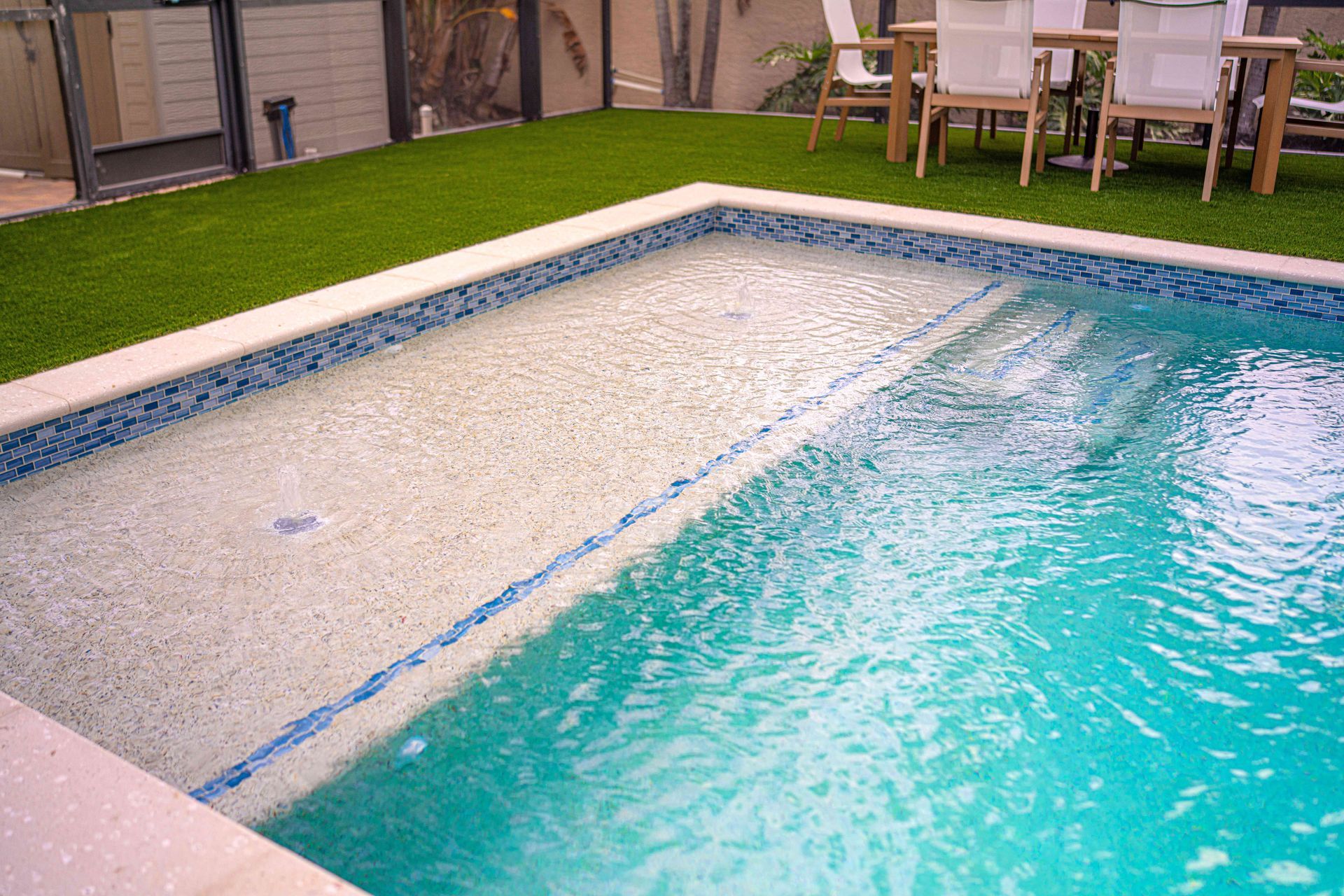 Backyard swimming pool with clear blue water, tiled edge, and lounge chairs on a patio nearby