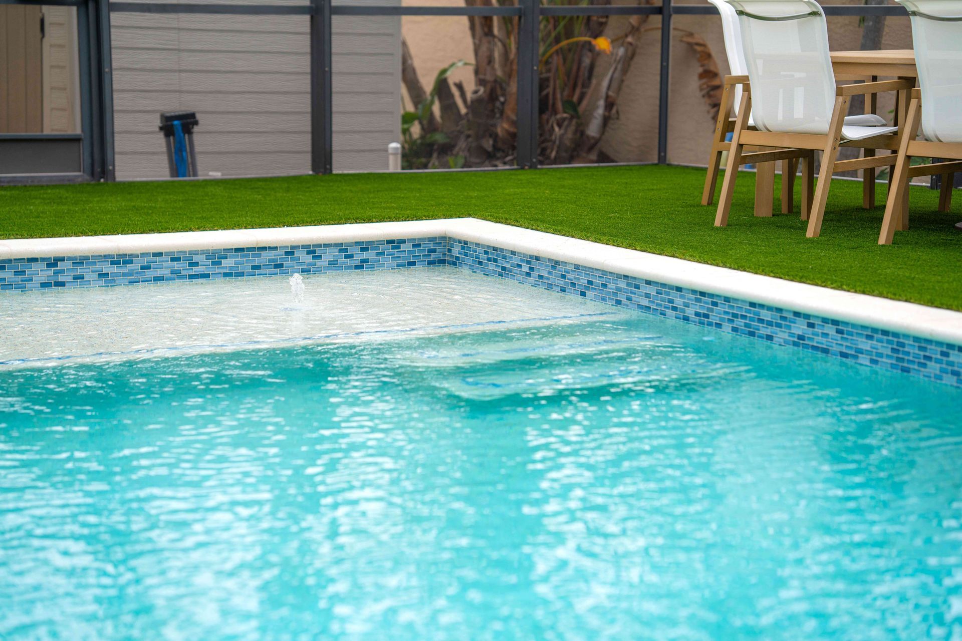 Outdoor swimming pool with turquoise water, white tiled edge, and patio chairs on a grassy deck