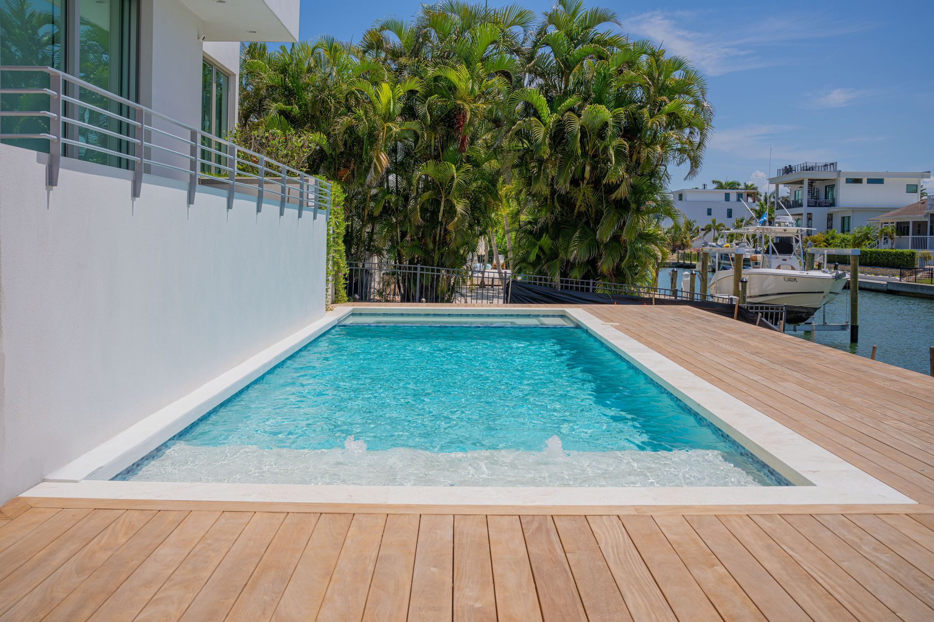 Modern backyard pool beside a white house, with a wooden deck and palm trees beyond the water.