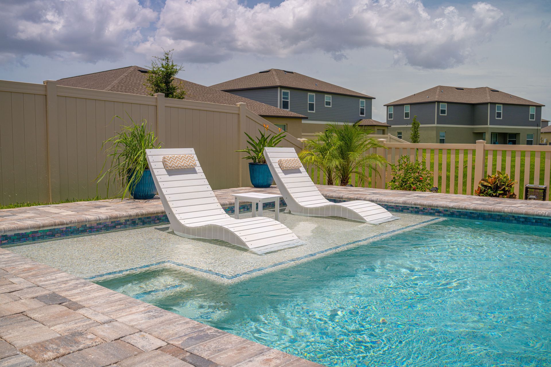 Backyard pool with two white lounge chairs on a stone deck under a cloudy sky