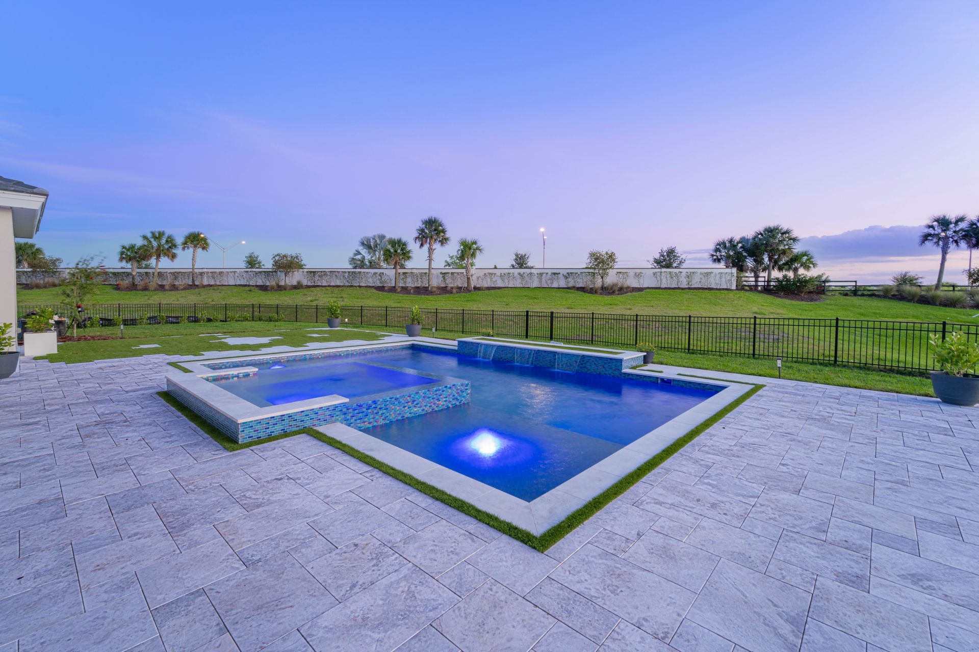 Modern backyard pool with blue lighting, stone patio, and ocean view at sunset