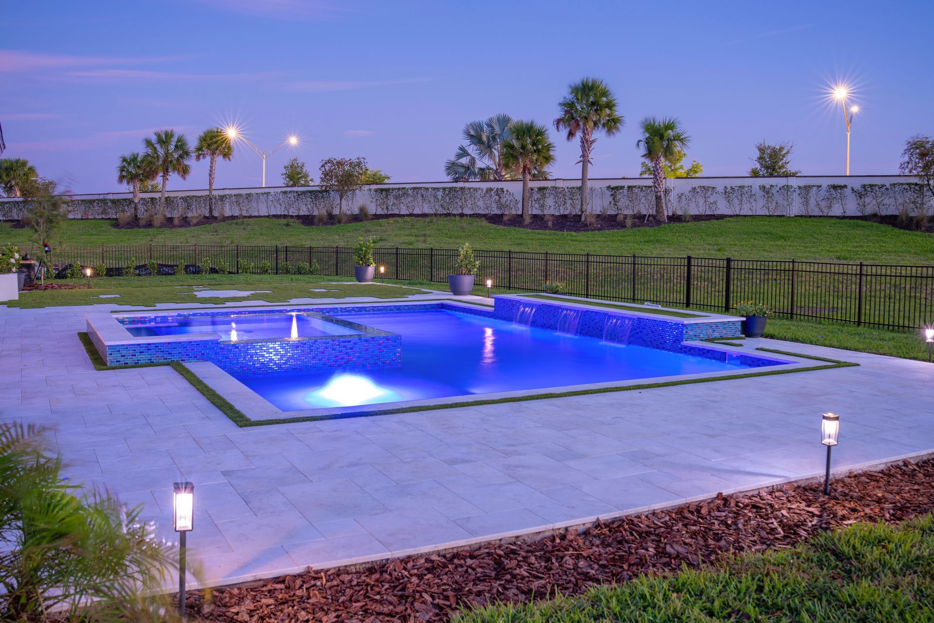 Blue-lit swimming pool beside a lawn and palm trees at dusk, with path lights around it