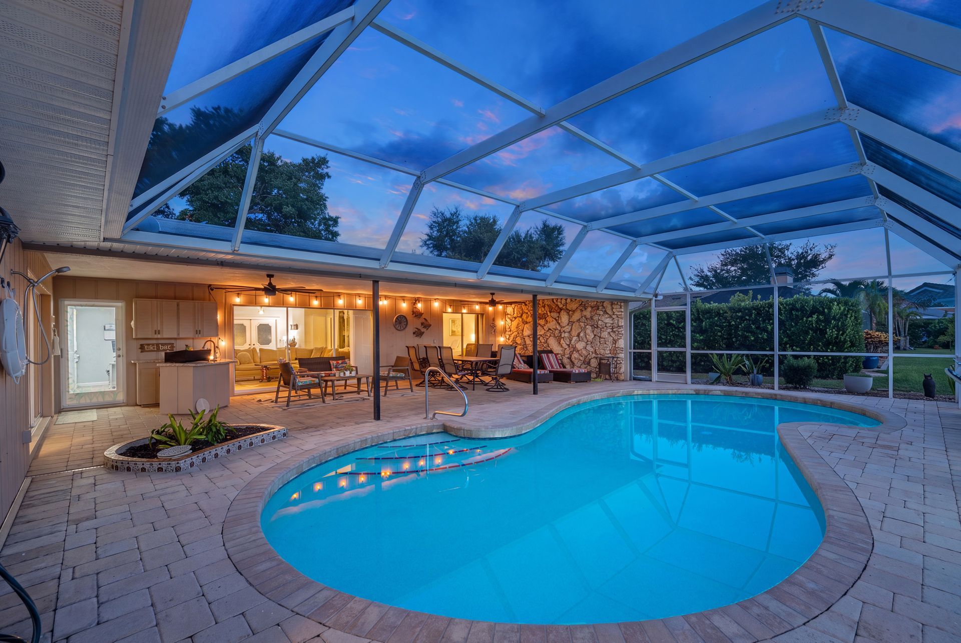 Indoor-outdoor patio with lit lounge area and curved blue swimming pool under a glass enclosure at dusk