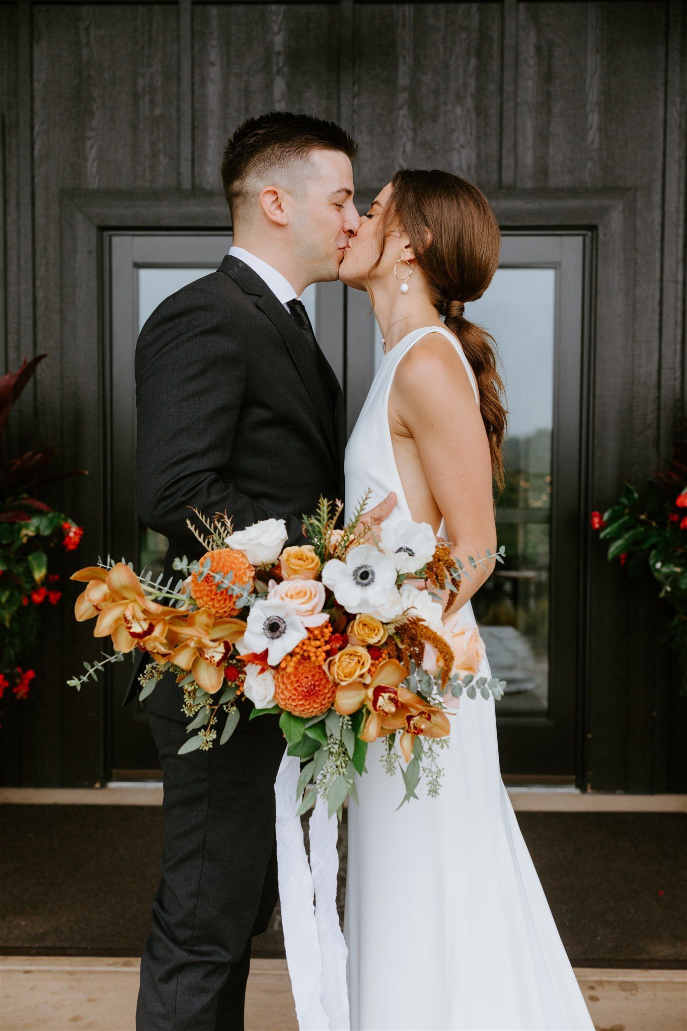 Bride and groom kissing, holding bouquet. Building background with floral arrangements.
