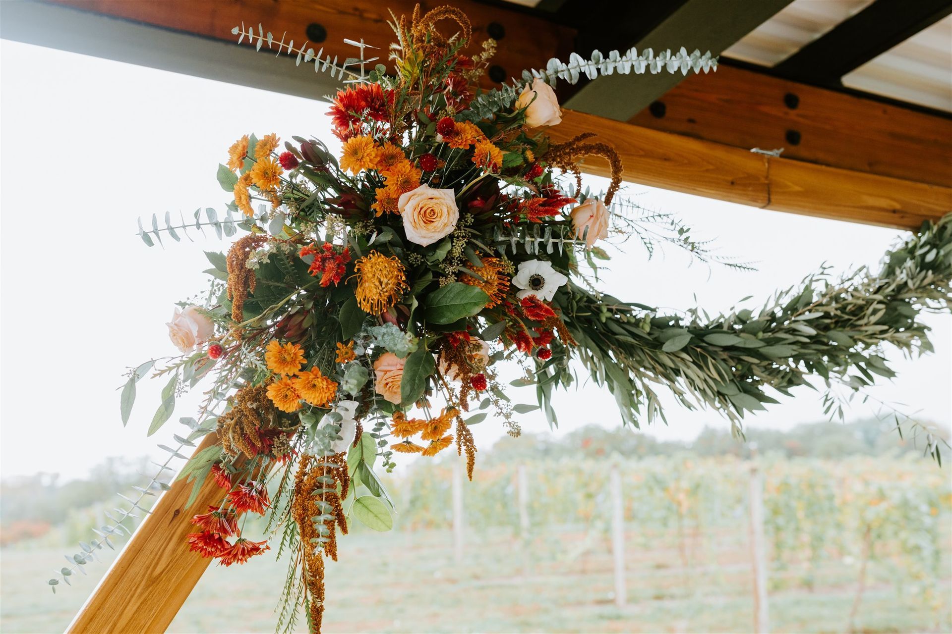 Floral arrangement with orange, red, and pink flowers and green foliage, against a vineyard background.