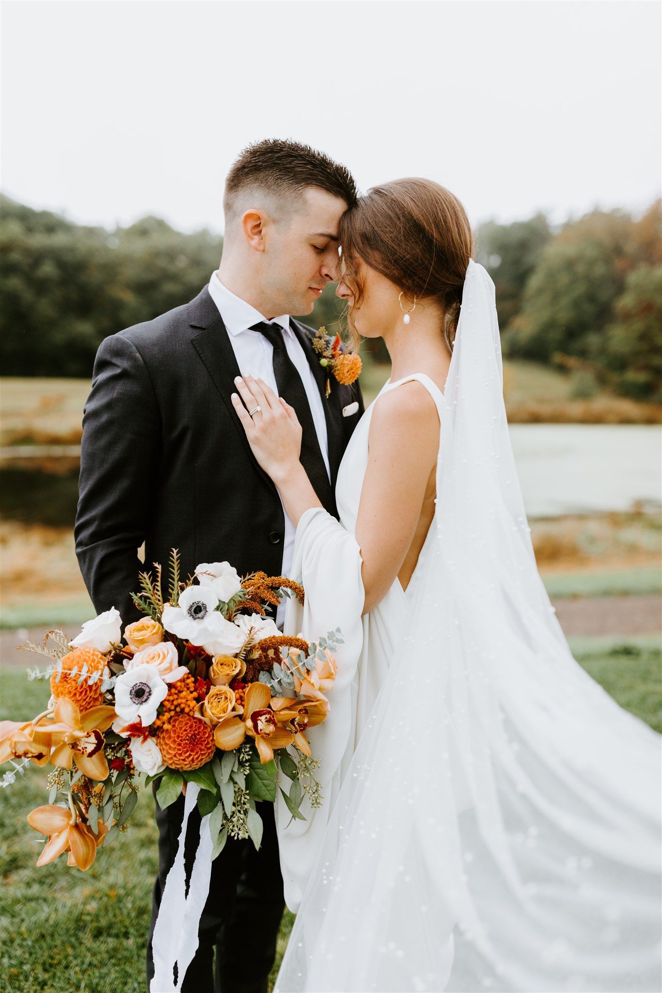 Bride and groom embracing, holding bouquet, outdoors near a pond. She wears white dress, veil; he wears suit.