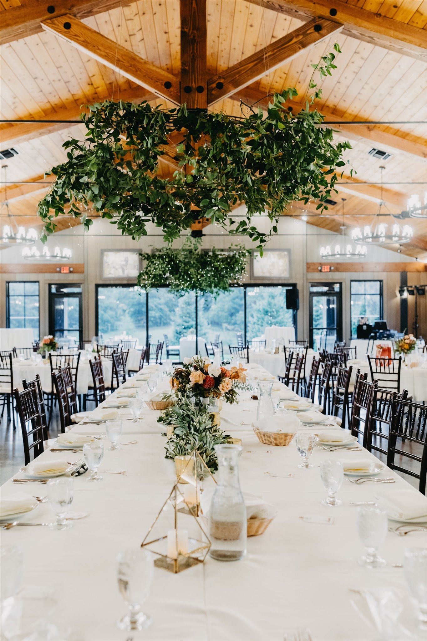Long banquet table set for a wedding reception in a wood-beamed hall; greenery hangs above.