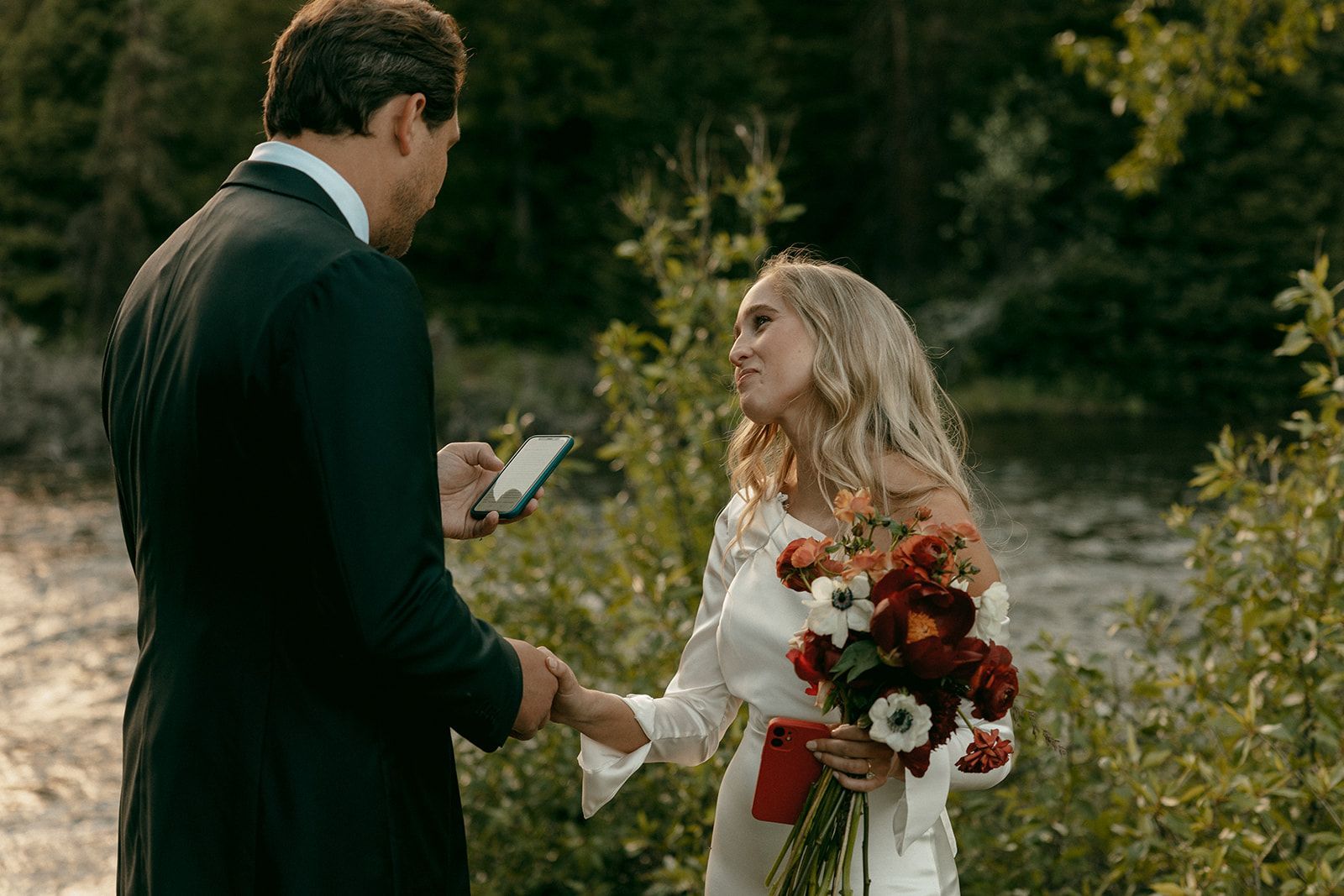 Couple in wedding attire exchanging vows by water, woman holds bouquet.