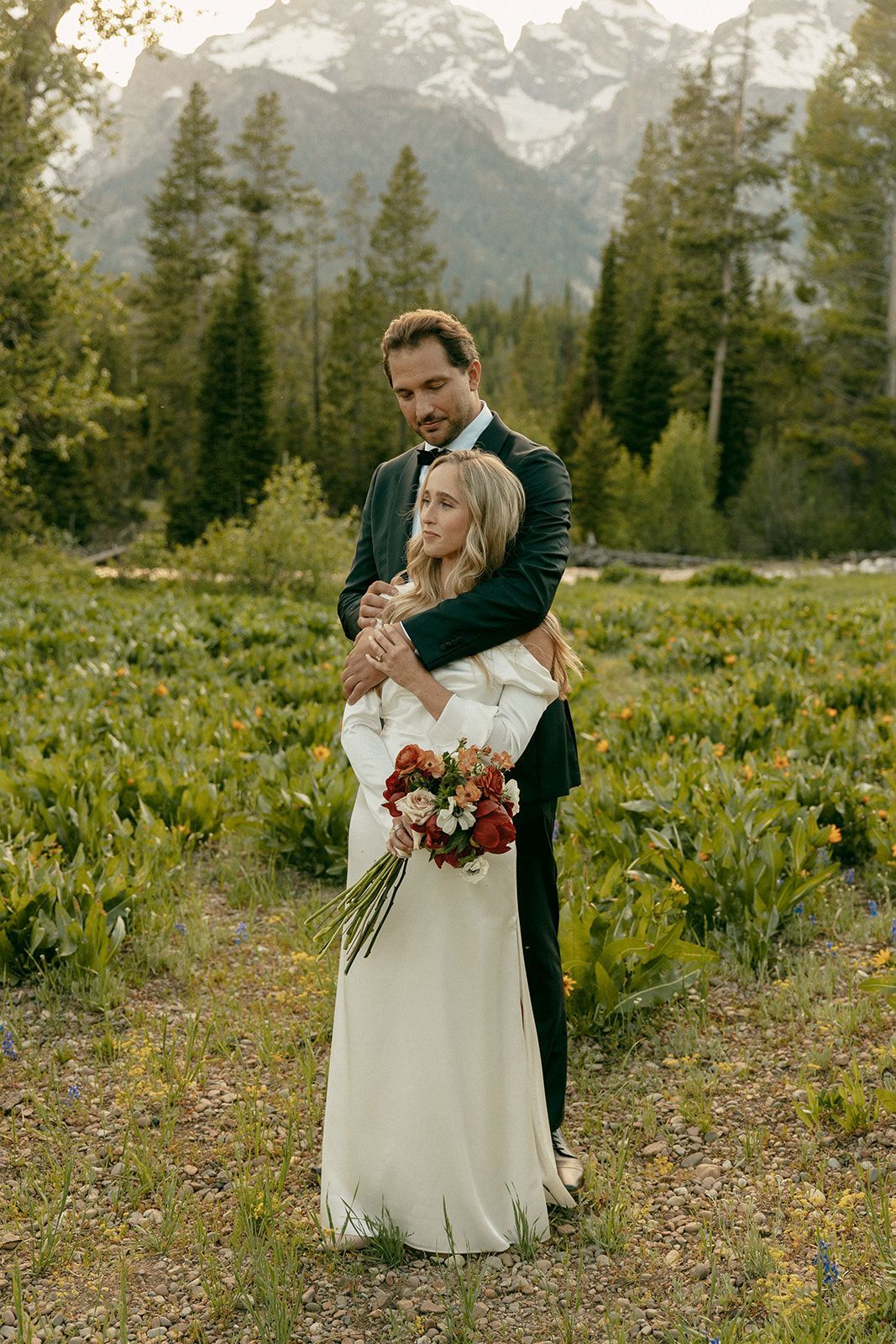 Couple embraces, wedding attire, holding bouquet, meadow, mountains in background.
