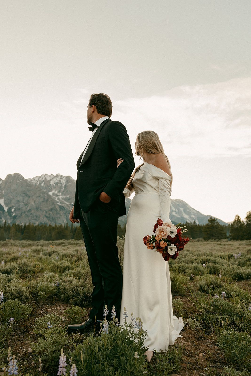 Bride and groom in formal attire stand in field, looking toward snow-capped mountains. The bride holds a bouquet.
