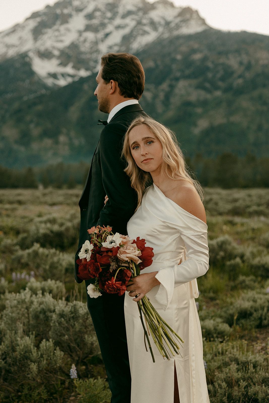 Bride in white dress, groom in tuxedo, mountain backdrop.