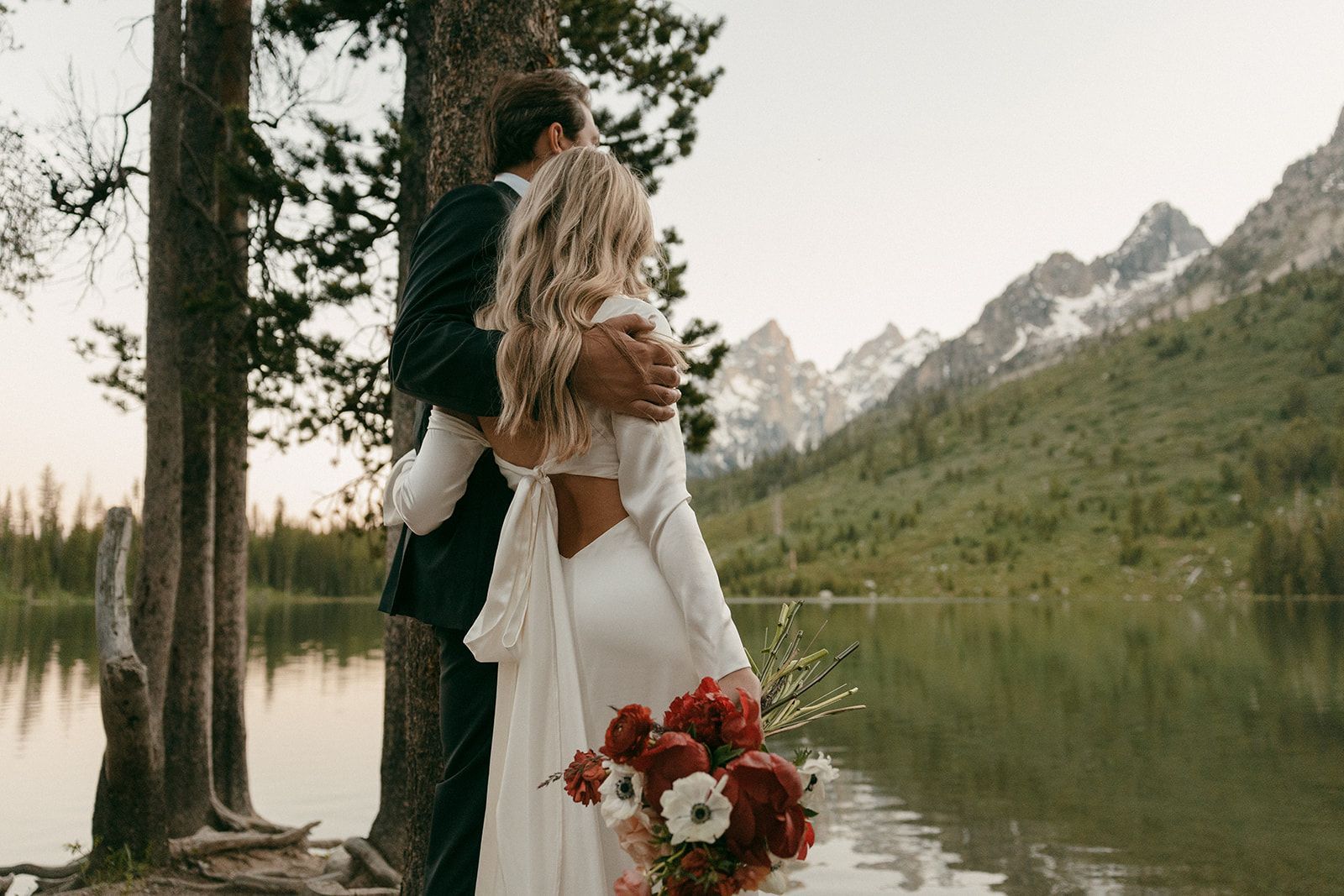 Couple embraces by a lake with mountains in the background; bride wears white dress with bouquet.